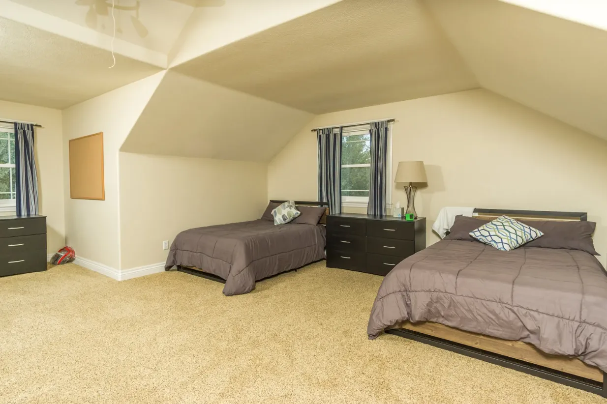 Shared bedroom with two twin beds, dark dressers, neutral walls, and windows with striped curtains under a sloped ceiling
