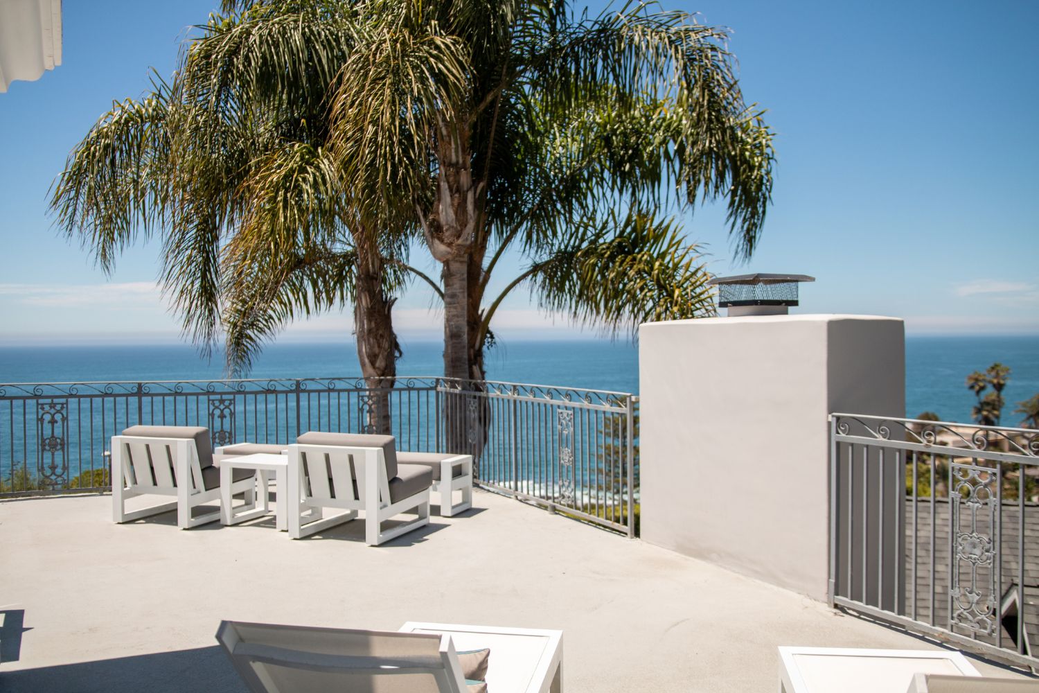 Stunning ocean-view patio with cozy seating, palm trees, and elegant railings, offering a serene and relaxing outdoor space under clear skies.