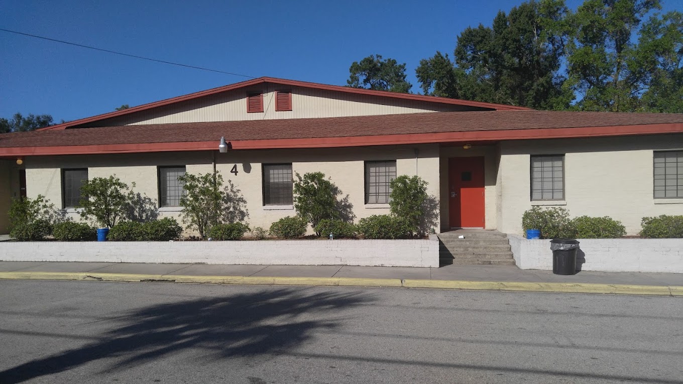 Street view of the facility with entrance. One-story beige building with red trim and shrubs