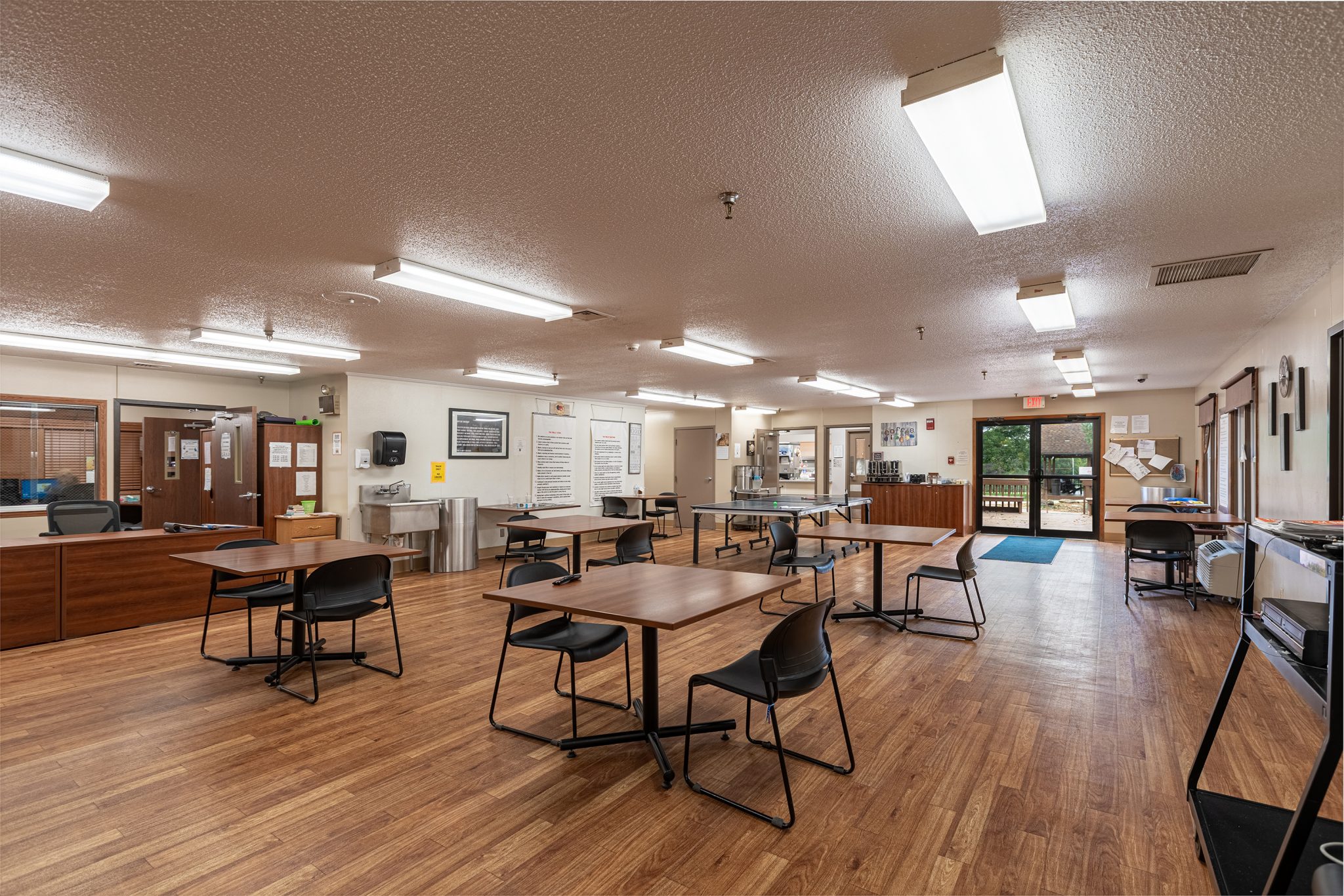 Spacious communal room with tables, chairs, and natural light from exterior doors.
