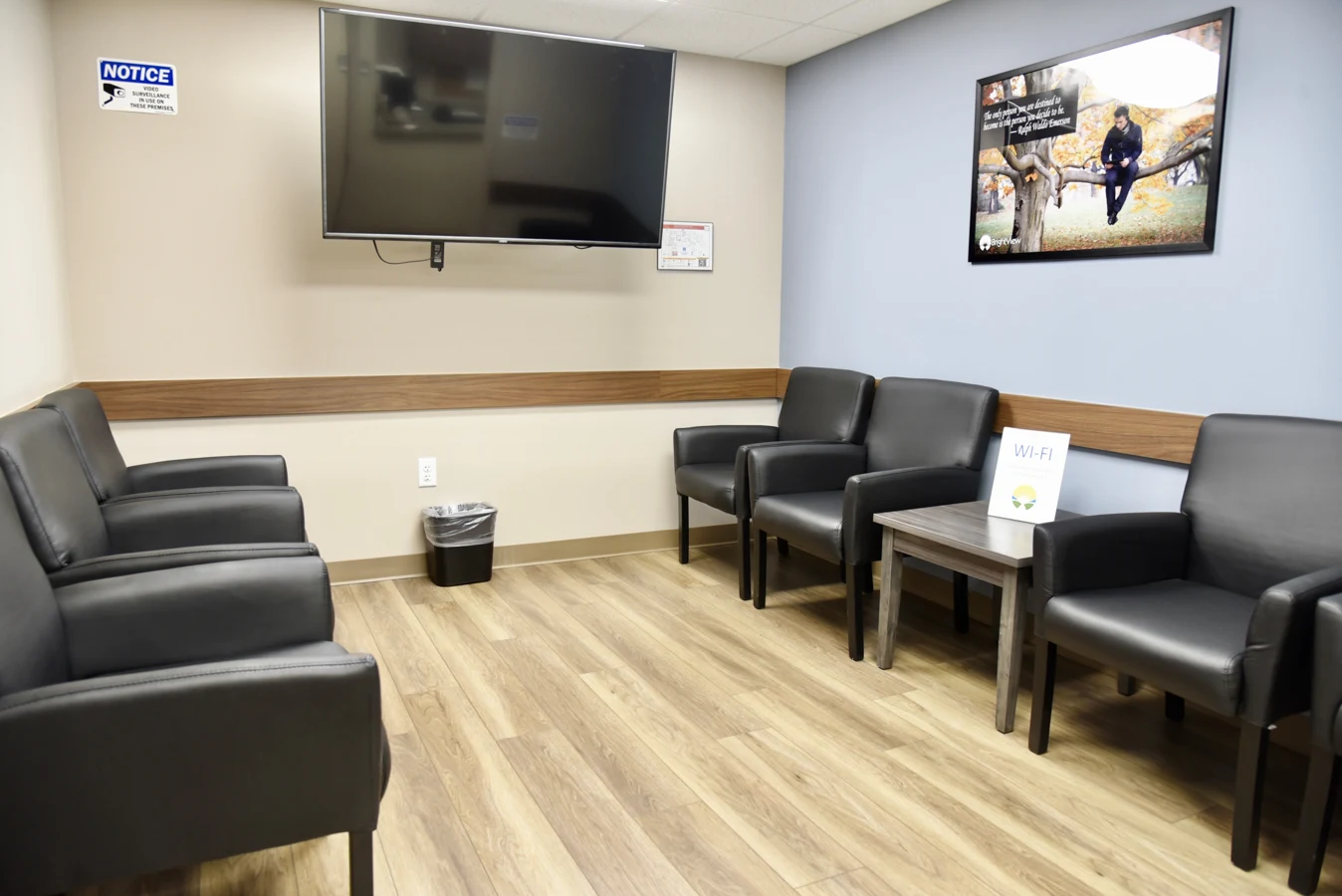 BrightView Health treatment waiting area with black cushioned chairs, wall-mounted television, Wi-Fi table sign, framed artwork, blue accent wall, and wood-look flooring