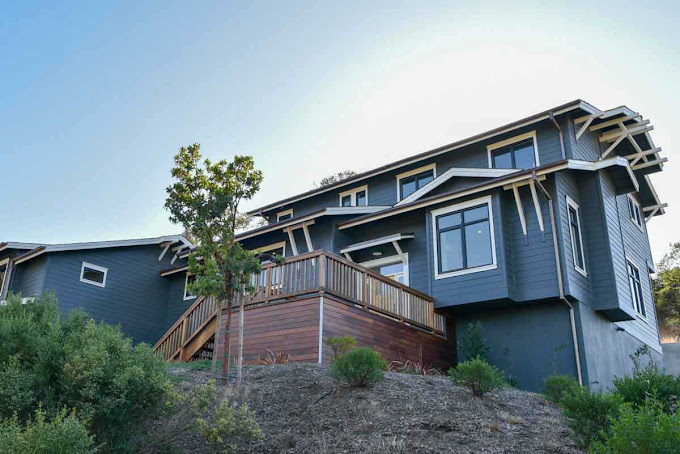 Side angle of gray building with wood deck, wooden stairs and shrubs