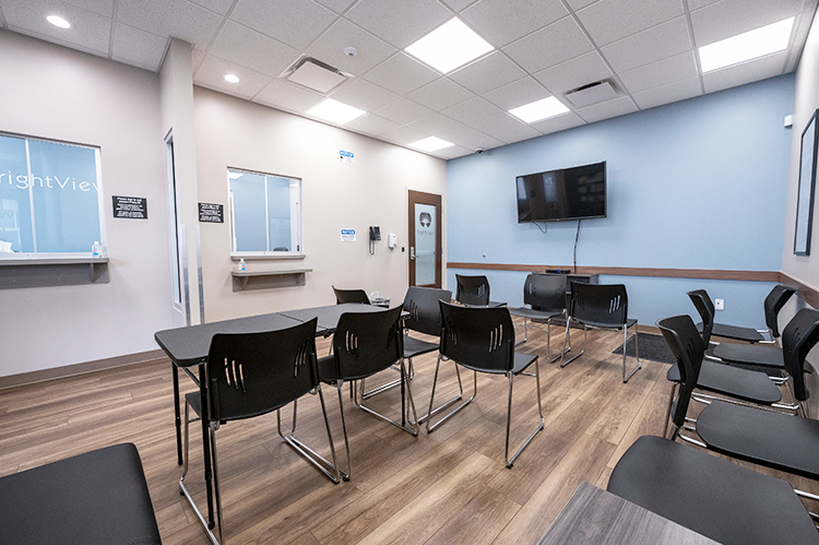 Group room with rows of black chairs and tables, wall-mounted television, reception windows, blue accent wall, and wood-look flooring
