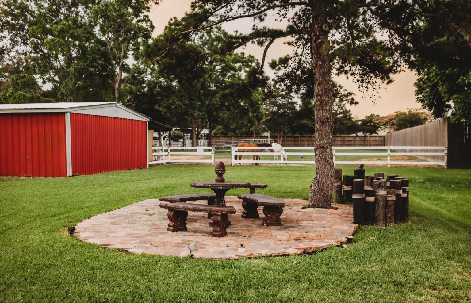 Outdoor stone patio with carved benches and table under a tree, near red barn and horse paddock