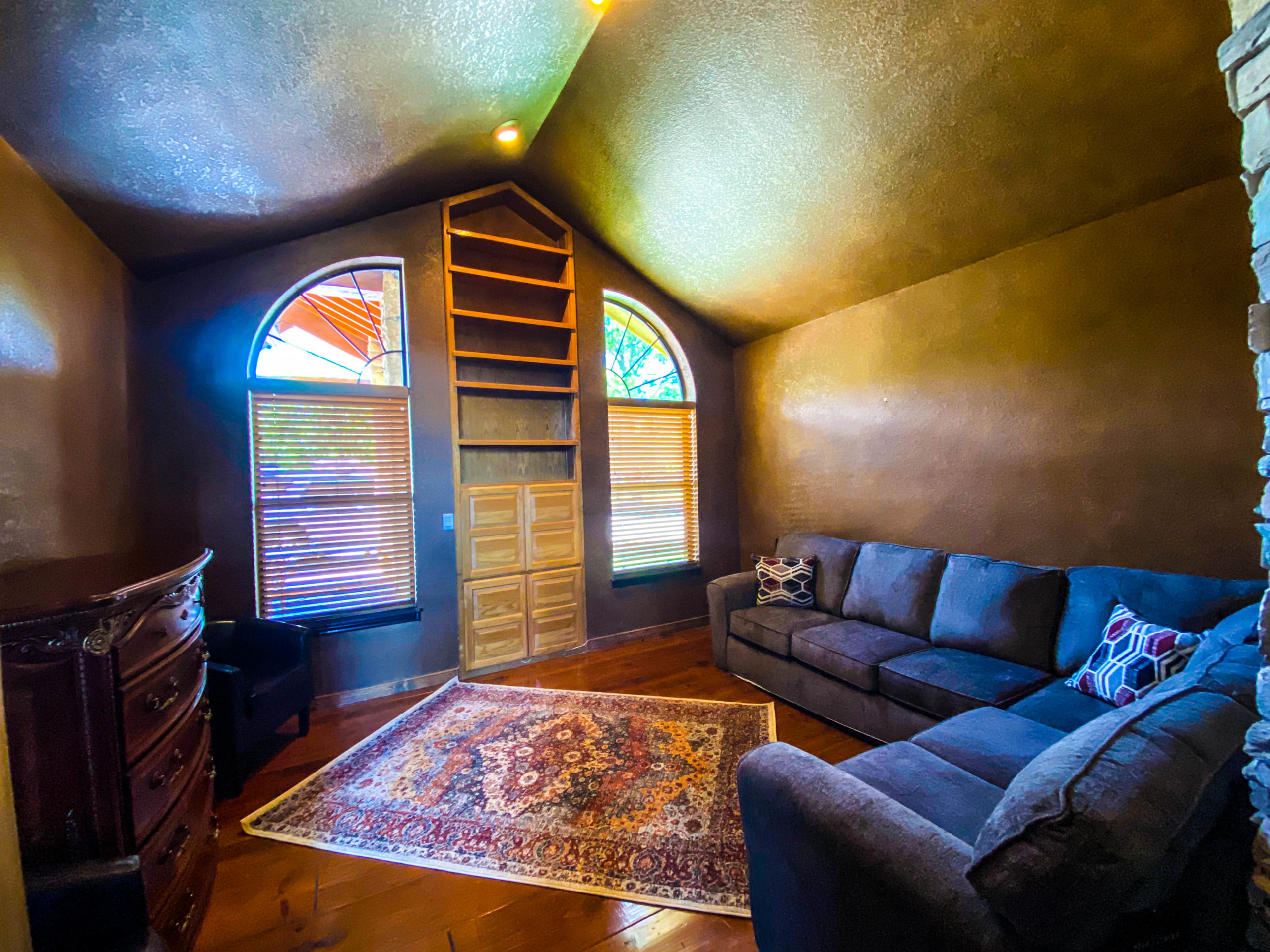 Cozy indoor room with arched windows, built-in shelving, a large sectional sofa, wooden flooring, and a decorative rug
