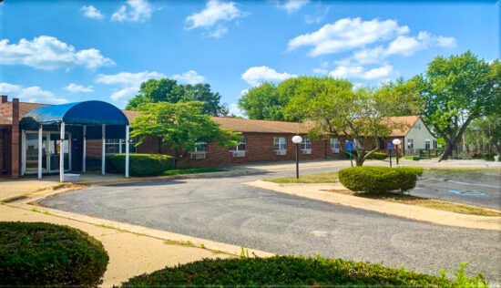 Brick facility with a blue awning, parking lot, and manicured trees