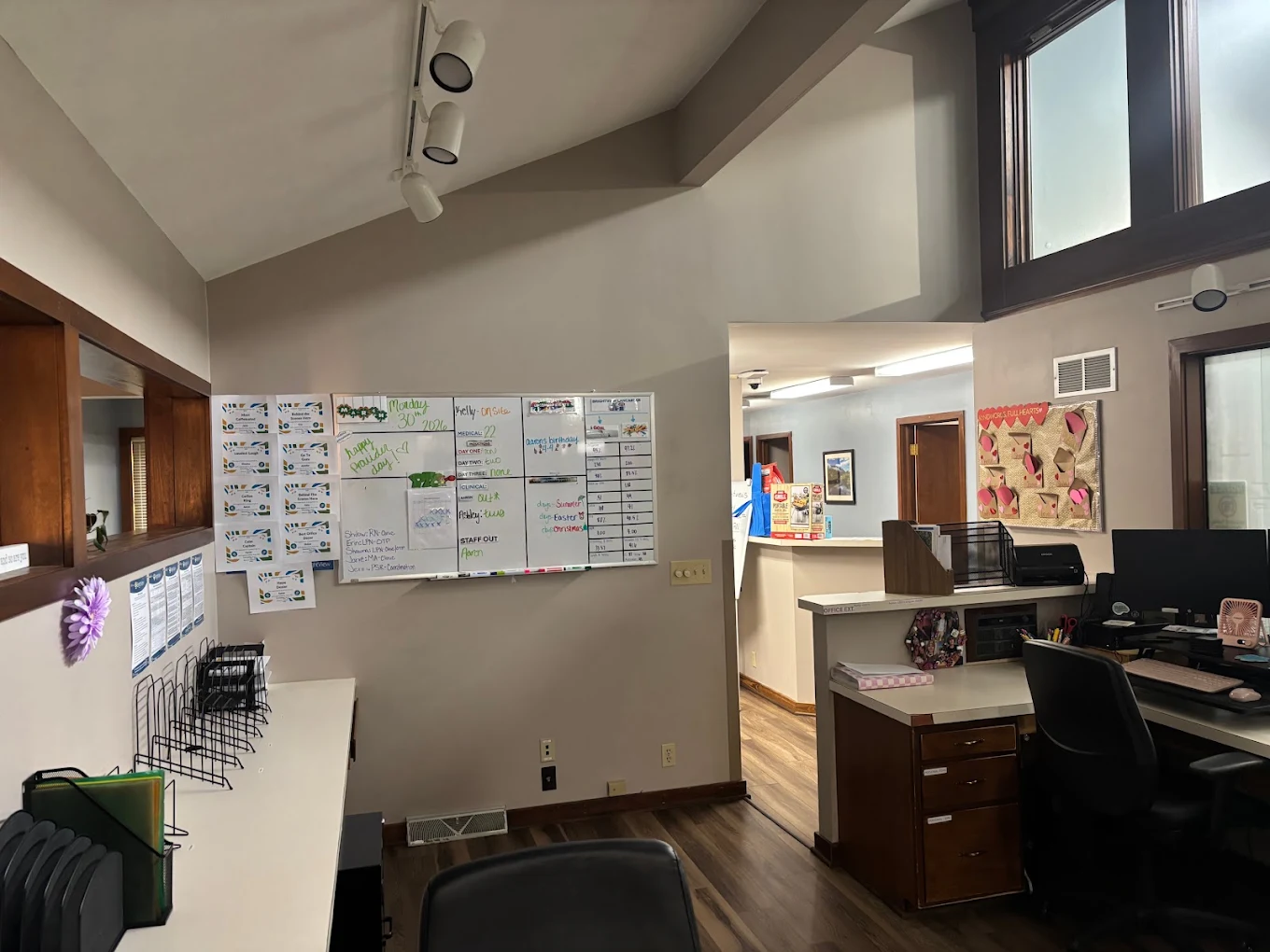 BrightView Health staff workstation with computer desks, office chairs, whiteboard, reception counter, wood-look flooring, and high windows