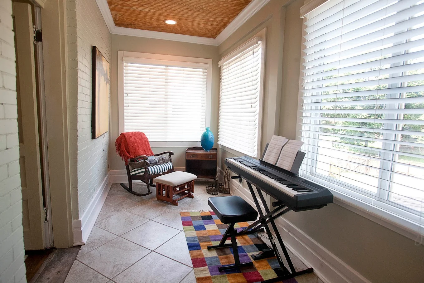 Bright room with electric piano, rocking chair, footrest, and windows with blinds