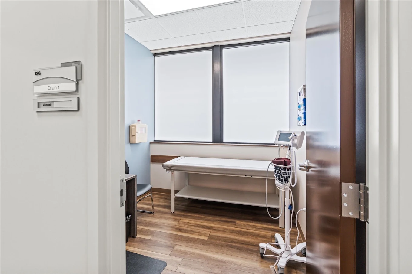 BrightView Health exam room with examination table, vital signs monitor, chair, wood-look flooring, and frosted windows