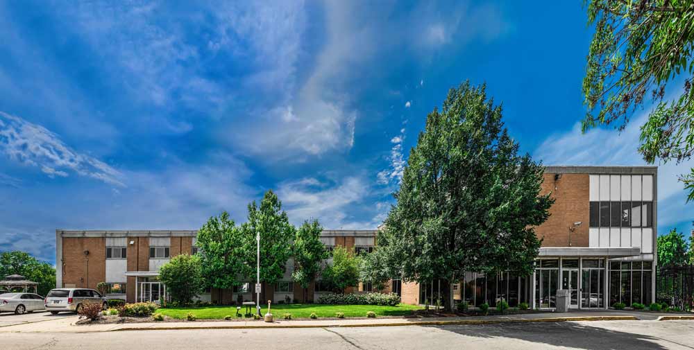 A modern facility with large windows, greenery, and spacious parking, providing a welcoming atmosphere under a bright blue sky.