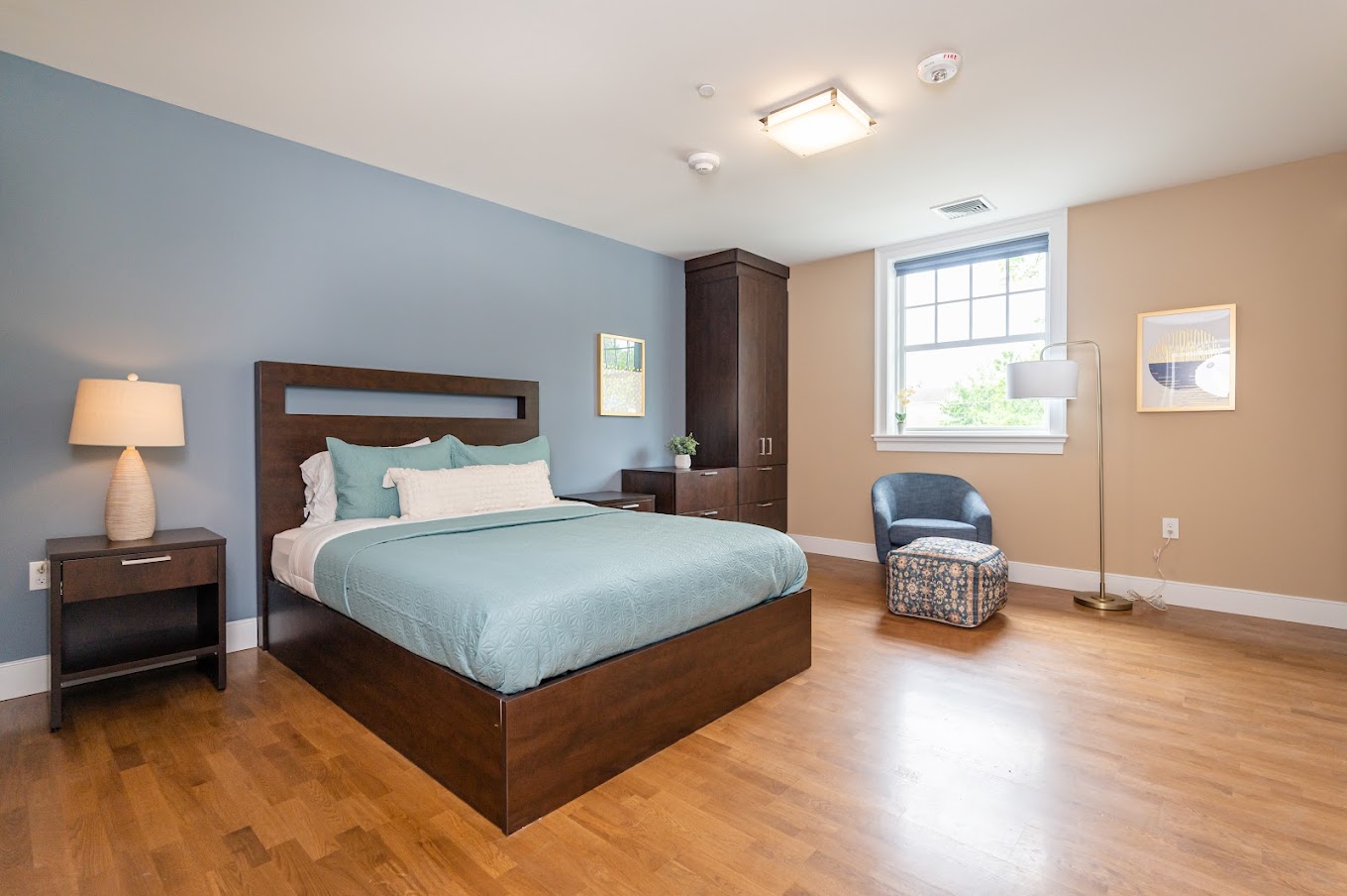  A contemporary and neatly arranged bedroom with a plush bed, wooden flooring, a bedside lamp, and a cozy armchair, creating a relaxing ambiance.