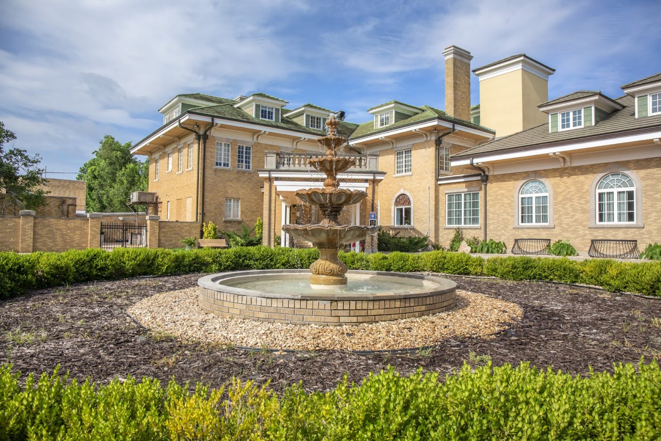 A landscaped circular driveway featuring a multi-tiered fountain in front of a historic-style brick building.