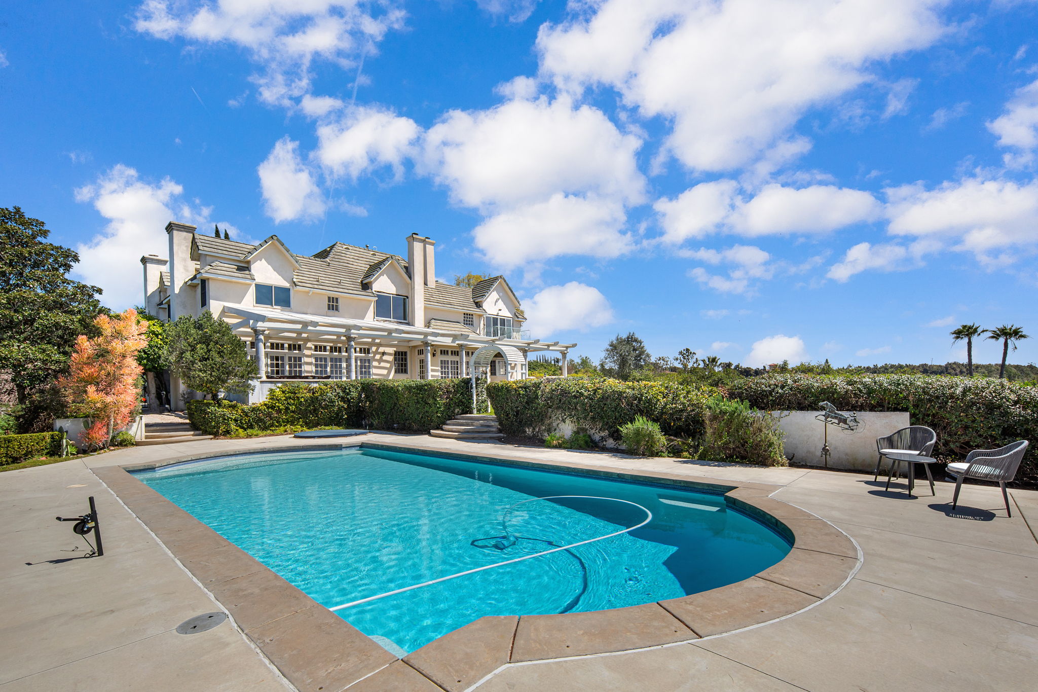 Pool area with lush landscaping and large house in background