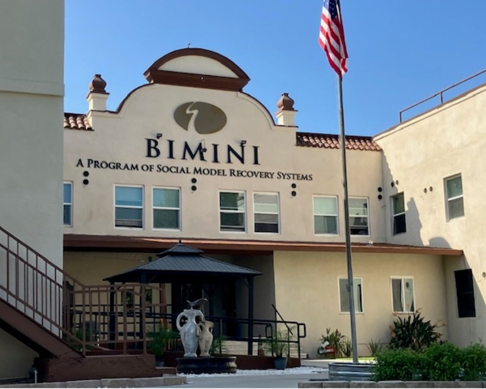 Front view of Bimini facility in Los Angeles with beige exterior, arched roofline, American flag, and fountain in front